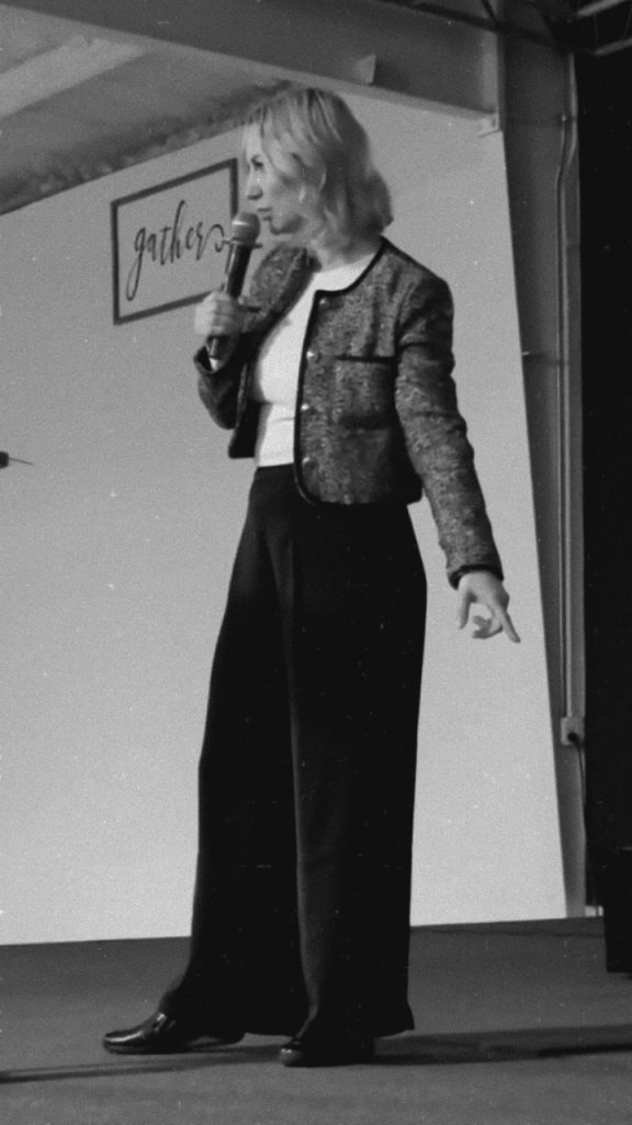 black and whoite photo of woman preaching, one hand on micriphone the other in a pointing direction to the floor. woman is onstage like on the church platform.